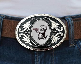May include: An oval-shaped silver and black metal belt buckle featuring an intricate filigree border and a central graphic of a small kitten holding a toy gun. The buckle is attached to a brown leather belt worn over dark blue denim jeans.