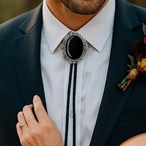 Op de afbeelding: Een bolo-das met een ovale, zilverkleurige metalen zetting met gedetailleerde filigraanranden rondom een gladde, effen zwarte steen. De hanger is bevestigd aan een dun, zwart gevlochten koord dat verticaal over een wit overhemd naar beneden hangt.