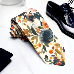 May include: A white tie with a floral pattern in shades of orange, green, and blue. The tie is made of a soft, silky fabric and has a subtle sheen.