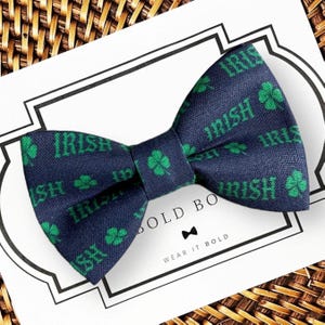 May include: A navy blue bow tie featuring a repeating pattern of green shamrocks and the word IRISH in green text. The bow tie is displayed on a white card with the text BOLD BOW and WEAR IT BOLD, along with a small bow tie icon.