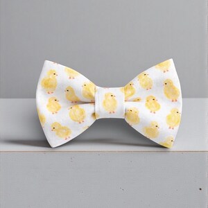 May include: A white bow tie featuring a repeating pattern of small, yellow, watercolor-style baby chicks. The fabric has a soft texture, and the bow tie is constructed with a central knot detail, showcasing a consistent print of fluffy birds.