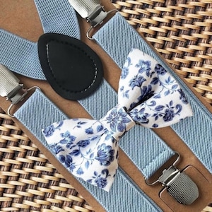 May include: Light blue suspenders with silver-tone metal clips and a black leather back patch. A matching bow tie with a white background and blue floral pattern completes the set. The accessories are displayed on a brown surface.