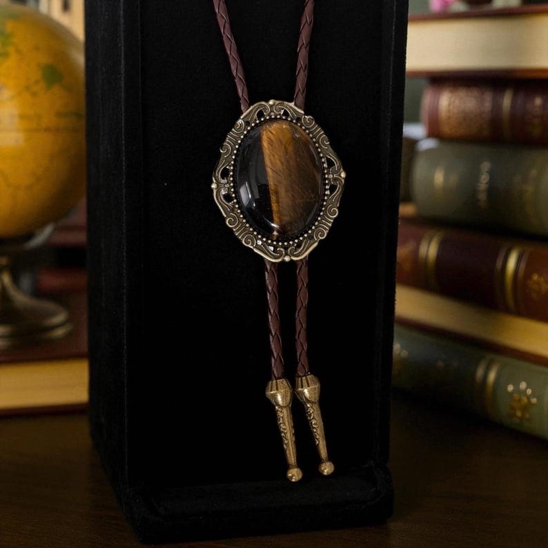 Puede incluir: Una corbata bolo con un colgante ovalado marr&oacute;n y dorado con una piedra ojo de tigre. El colgante tiene un borde met&aacute;lico ornamentado de estilo antiguo. La corbata tiene un cord&oacute;n trenzado marr&oacute;n con puntas met&aacute;licas decorativas.