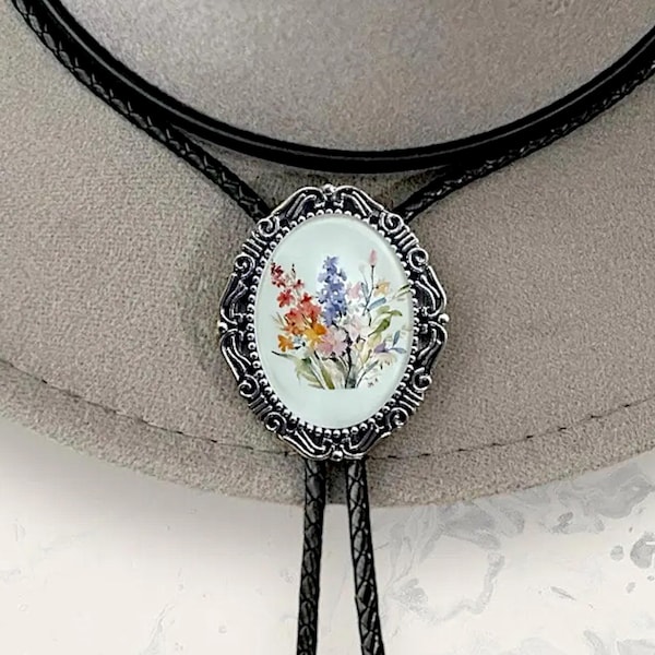 Puede incluir: Una corbata bolo con un engaste ovalado de metal plateado con bordes de filigrana intrincados. El centro muestra una ilustración colorida de un ramo de flores estilo acuarela sobre fondo blanco, unida a un cordón trenzado negro.