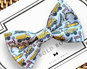 May include: A light blue bow tie featuring a repeating pattern of yellow construction vehicles, including dump trucks and excavators, along with red and white striped road barriers and traffic cones. The bow tie is displayed on a white card.