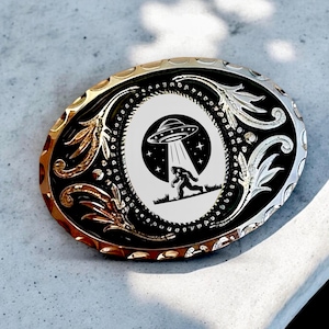 Könnte beinhalten: Eine silberne und schwarze Gürtelschnalle mit einem weißen ovalen Design, das eine schwarze Silhouette eines Bigfoots zeigt, der von einem UFO weggeht, aus dem ein Lichtstrahl nach unten scheint. Die Schnalle hat ein detailliertes Blumenmuster am Rand.