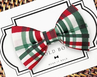 May include: A fabric bow tie featuring a plaid pattern in red, dark green, and white. The bow tie is attached to a white display card with the text "BOLD BOW" and "WEAR IT BOLD" printed in black, along with a small black bow tie icon.