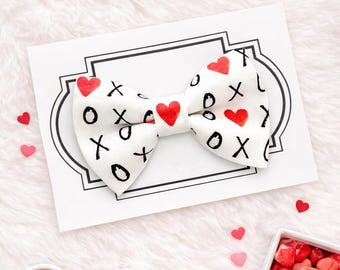 May include: A white bow tie featuring a repeating pattern of black handwritten X and O symbols interspersed with small red hearts. The bow tie is centered on a rectangular white card with a black decorative border frame design.