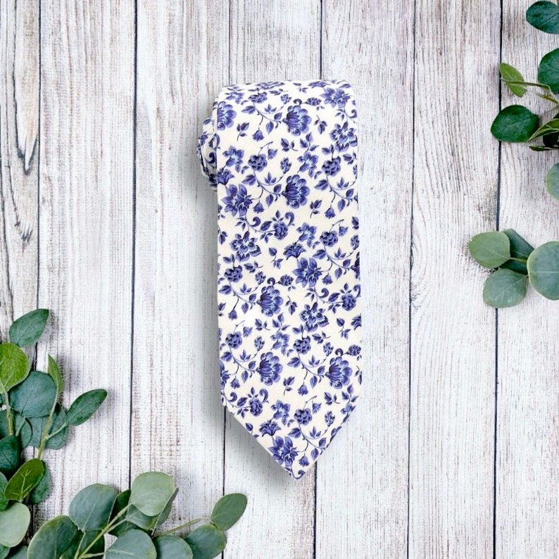 Op de afbeelding: Een opgerolde stropdas met een bloemenpatroon. De stropdas heeft een cr&egrave;mekleurige achtergrond, versierd met een herhalend patroon van blauwe bloemen en bladeren. De stof voelt zacht aan, geschikt voor formele of semi-formele gelegenheden. De stropdas is ongeveer 140 cm lang.