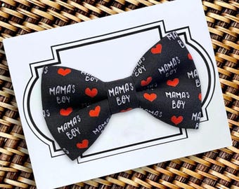 May include: A black fabric bow tie featuring a repeating pattern of small red hearts and the white text "MAMA'S BOY" printed across the surface. The bow tie is attached to a white rectangular display card with a black border outline.