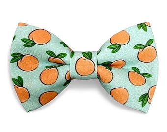 May include: A bow tie featuring a repeating pattern of orange peaches with green leaves on a light mint green background. The fabric has a visible woven texture, and the bow tie is constructed with a central knot piece connecting two wings.