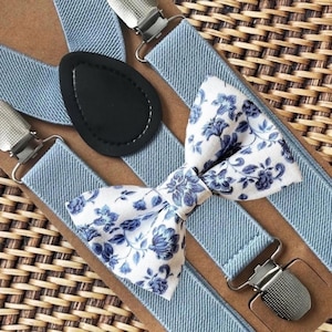 May include: A set of light blue elastic suspenders featuring silver metal clips and a black leather-like teardrop-shaped back patch, paired with a matching white bow tie decorated with a blue floral pattern on a brown cardboard backing.