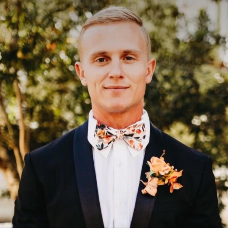 May include: A man wearing a black suit jacket with a white shirt and a floral bow tie. He is wearing a boutonniere with a peach-colored flower.
