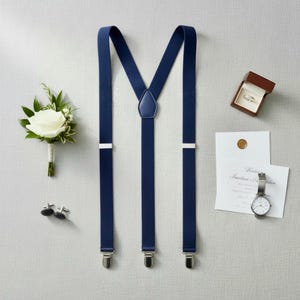 Puede incluir: Tirantes azul marino con cierres plateados, un prendido de rosa blanca, gemelos, un reloj, un anillo de bodas en una caja y una invitación de boda. Los tirantes tienen un parche de cuero.