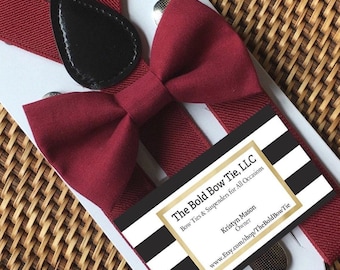 May include: A set of burgundy suspenders and a matching bow tie featuring a black leather-like accent piece. The set is displayed on a white card with a black and white striped label that reads The Bold Bow Tie, LLC, Bow Ties & Suspenders for All Occasions.
