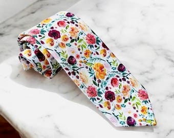Puede incluir: Una corbata con fondo blanco y un vibrante estampado floral. El diseño incluye racimos de flores en tonos burdeos intenso, amarillo brillante, rosa suave y follaje verde, dispuestos en un estampado botánico repetitivo.
