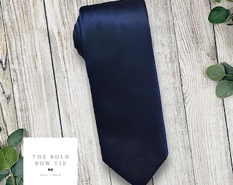 Puede incluir: Una corbata de color azul marino sólido con un acabado suave y satinado. Una etiqueta cuadrada blanca se encuentra en la esquina inferior izquierda con el texto THE BOLD BOW TIE y la frase WEAR IT BOLD junto a un pequeño icono de pajarita.