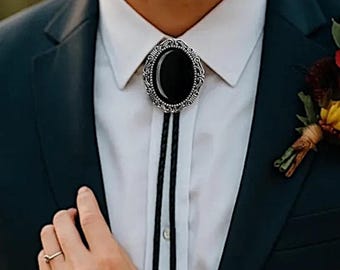 May include: A bolo tie featuring an oval-shaped black stone set in a decorative silver-toned metal frame with intricate scrollwork details, attached to a thin black cord worn over a crisp white button-down shirt and a dark suit jacket.