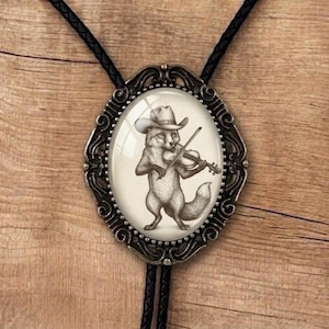 May include: Oval bolo tie with a silver-coloured filigree frame. The focal point is a drawing of a fox wearing a cowboy hat and playing a violin. The tie has a black leather cord.