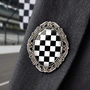 May include: Silver-toned oval brooch with a black and white checkered design. The brooch is set against a dark gray fabric, with a racing flag visible in the background. The brooch has an ornate, filigree-style frame.