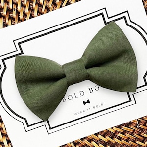 Könnte beinhalten: Eine olivgrüne Fliege aus Stoff, zentriert auf einer weißen rechteckigen Präsentationskarte. Die Karte hat einen schwarzen dekorativen Rand und den aufgedruckten Text BOLD BOWS mit einem kleinen Fliegen-Symbol und dem Schriftzug WEAR IT BOLD.