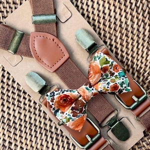 May include: A set of brown elastic suspenders featuring tan leather accents and antique brass-toned metal clips, paired with a matching bow tie that displays a floral pattern in shades of orange, green, and white on a light background.