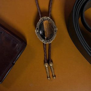 Puede incluir: Una corbata bolo con un cord&oacute;n trenzado marr&oacute;n y un colgante ovalado decorativo. El colgante presenta una piedra ojo de tigre engastada en un marco de metal ornamentado de estilo antiguo. Tambi&eacute;n se ven una cartera de cuero marr&oacute;n y un cintur&oacute;n negro.