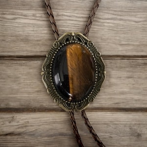 Puede incluir: Corbata bolo ovalada con una piedra ojo de tigre engastada en un marco ornamentado de color lat&oacute;n antiguo. La piedra es marr&oacute;n y negra. El cord&oacute;n de cuero trenzado es de color marr&oacute;n oscuro. Un accesorio western cl&aacute;sico.