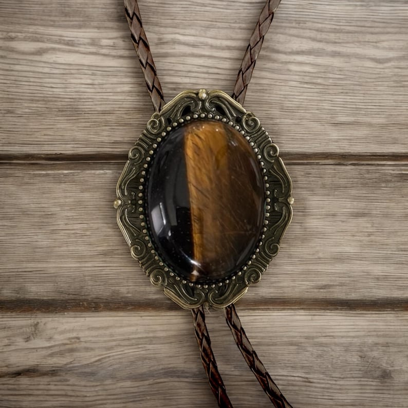 Puede incluir: Corbata bolo ovalada con una piedra ojo de tigre engastada en un marco ornamentado de color lat&oacute;n antiguo. La piedra es marr&oacute;n y negra. El cord&oacute;n de cuero trenzado es de color marr&oacute;n oscuro. Un accesorio western cl&aacute;sico.