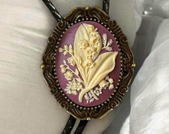 May include: A bolo tie featuring an oval-shaped cameo pendant with a deep purple background and a raised, cream-colored relief of lily of the valley flowers. The pendant is encased in an ornate, antique-style bronze metal frame.
