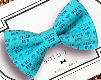 May include: A bright turquoise bow tie featuring a repeating pattern of dark blue palm trees and the text BEACH BUM. The bow tie is displayed on a white card with the text BOLD BOW, a small bow tie icon, and the phrase WEAR IT BOLD.