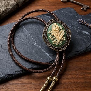 May include: A bolo tie featuring a dark brown braided leather cord with ornate metal tips. The central oval pendant has an antique bronze-toned frame surrounding a green inlay with a raised, cream-coloured floral relief of lily of the valley.