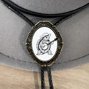 Puede incluir: Un corbatín bolo con un cordón de cuero marrón y un colgante ovalado de bronce con una ilustración en blanco y negro de una tortuga con un sombrero de vaquero tocando un banjo.
