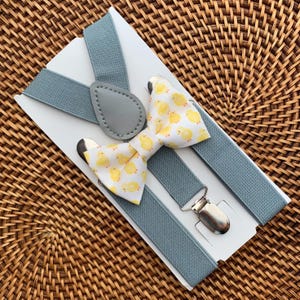 May include: A set consisting of a bow tie and suspenders. The bow tie is white with a repeating pattern of small yellow chicks. The suspenders are a solid dusty blue color and feature silver-toned metal clips and a gray faux leather patch.