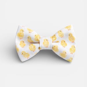 May include: A white bow tie featuring a repeating pattern of small, yellow, watercolor-style baby chicks. The bow tie has a central fabric knot and a classic butterfly shape, with the chick illustrations distributed evenly across the fabric surface.