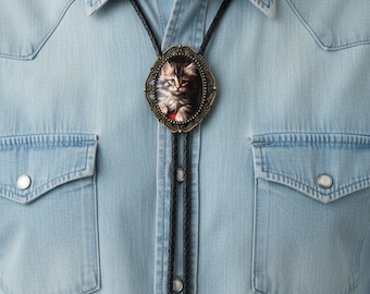 May include: A bolo tie featuring an oval-shaped antique bronze frame containing a detailed image of a fluffy kitten. The tie consists of a black braided cord with matching bronze-toned metal tips, worn over a light blue denim button-down shirt.