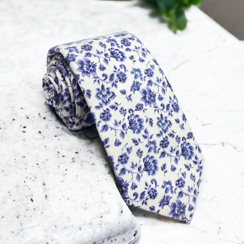 Op de afbeelding: Gerolde stropdas met een cr&egrave;mekleurige achtergrond en een herhaald patroon van blauwe bloemmotieven. De stropdas is gemaakt van een zachte stof en is geschikt voor formele of informele gelegenheden.