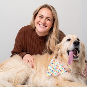 May include: A woman with long blonde hair smiles at the camera while sitting on the floor with a golden retriever dog. The dog is wearing a colorful bandana with the text "It's My Birthday" printed on it.