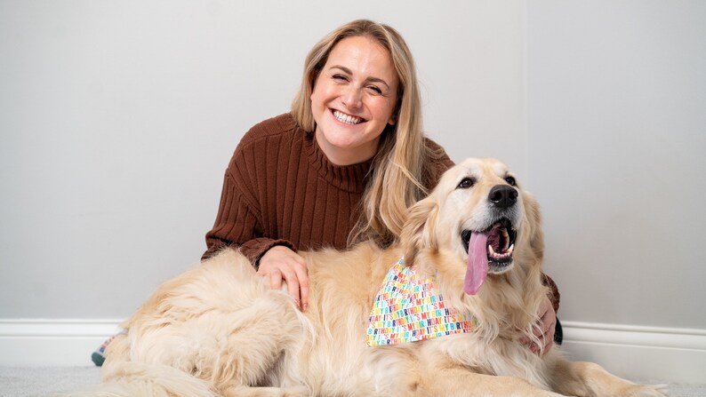 May include: A woman with long blonde hair smiles at the camera while sitting on the floor with a golden retriever dog. The dog is wearing a colorful bandana with the text "It's My Birthday" printed on it.