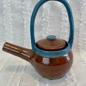 May include: A brown ceramic teapot with a blue handle and lid. The teapot has a rounded body and a spout on the side.