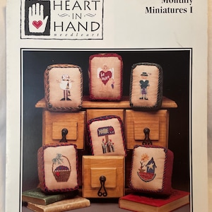 May include: A cover for "Heart in Hand Needleart" Monthly Miniatures I. The image features a wooden cabinet with several small, embroidered pillows. Each pillow has a different design, including a snowman, a heart, and a basket.