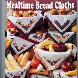 May include: A cover of a leaflet titled "Mealtime Bread Cloths" featuring baskets of bread and fruit. The baskets are lined with white cloths, each with a different embroidered design. The cover has the text "By Lois Winston".