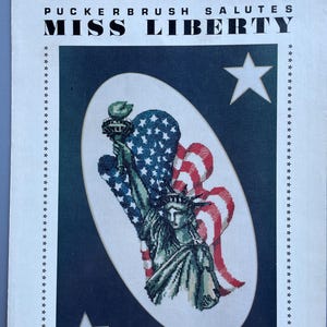 May include: A cross-stitch pattern featuring the Statue of Liberty with an American flag design. The text "PUCKERBRUSH SALUTES MISS LIBERTY" is at the top. The image is set against a dark blue background with white stars.