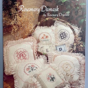 May include: A collection of embroidered pillows with floral and initial designs, displayed in a basket. The pillows feature white fabric with pink and green embroidery, and lace trim. The cover of the book reads "Rosemary Damask by Rosemary Drysdale".