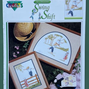 May include: A cross-stitch pattern titled "Swing Shift" by Steve Polomchak. The image features two framed cross-stitch samplers with pastoral scenes, a straw hat, and a small doll. The text reads "The carefree days of summer, when life was much simpler, is captured in Steve Polomchak's 'Swing Shift'."