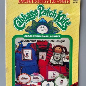May include: A vintage Cabbage Patch Kids cross stitch pattern book. The cover is yellow with the Cabbage Patch Kids logo and illustrations of cross-stitched items like a backpack, overalls, and bibs. The book is titled "Cross Stitch Small & Sweet" and features 10 designs.