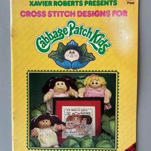 May include: A vintage cross-stitch design book titled "Xavier Roberts Presents Cross Stitch Designs For Cabbage Patch Kids." The cover is yellow with the Cabbage Patch Kids logo and images of the dolls. The book includes the text "All Babies are Special."