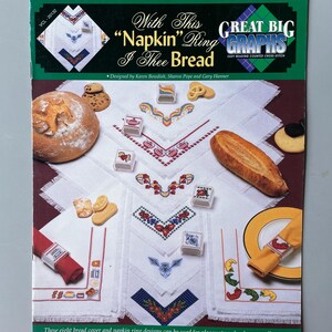 May include: A book cover with a white background and a colorful design of bread, napkin rings, and a title that reads "With This "Napkin" Ring I Thee Bread" and "Great Big Graphs Easy Reading Counted Cross Stitch".