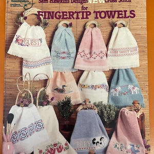 May include: A book cover with the title "Sam Hawkins Designs NEW Cross Stitch for FINGERTIP TOWELS". The cover features a variety of colorful cross-stitch designs on white towels, including a guest towel, a home sweet home towel, and a baby towel.