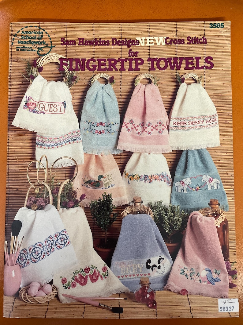 May include: A book cover with the title "Sam Hawkins Designs NEW Cross Stitch for FINGERTIP TOWELS". The cover features a variety of colorful cross-stitch designs on white towels, including a guest towel, a home sweet home towel, and a baby towel.